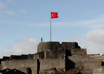 Açık mavi gökyüzü altında dalgalanan Türk bayrağı ve tarihi kale duvarları.