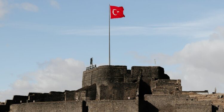 Açık mavi gökyüzü altında dalgalanan Türk bayrağı ve tarihi kale duvarları.