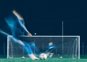 Gece saatlerinde futbol sahasında kaleye doğru şut çeken futbolcu.