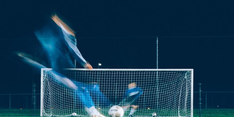 Gece saatlerinde futbol sahasında kaleye doğru şut çeken futbolcu.