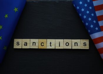 Siyah zemin üzerinde ahşap harflerle yazılmış sanctions kelimesi ve iki ülke bayrağı