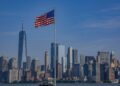 New York silüeti önünde dalgalanan Amerikan bayrağı ve denizdeki feribot.