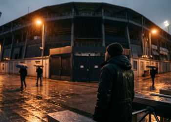 Yağmurlu bir akşamüstü stadyum önünde bekleyen kasketli ve montlu bir adam.
