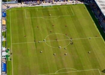 Havadan çekilmiş yeşil çim saha üzerinde futbol maçı oynayan sporcular.