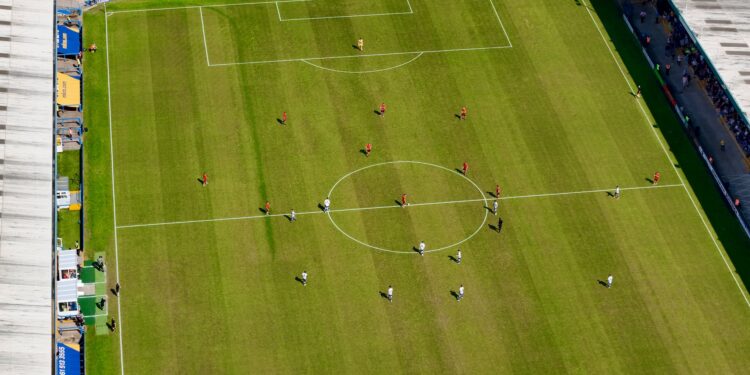 Havadan çekilmiş yeşil çim saha üzerinde futbol maçı oynayan sporcular.