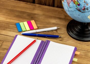 Ahşap masa üzerinde açık defter, tükenmez kalem, renkli yapışkan notlar ve küre.