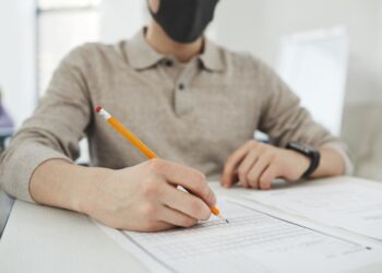 Aday, masada oturmuş maskeyle test kağıdını kurşun kalemle dolduruyor.