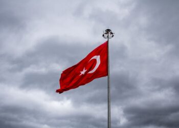 Bulutlu gökyüzü altında dalgalanan büyük Türk bayrağı ve aydınlatma direği.