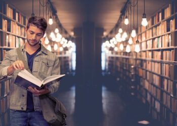 Kütüphanede kitap rafları arasında kitap inceleyen genç bir öğrenci.